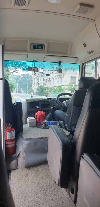 The image shows the interior of a bus with the view towards the windshield and dashboard. A digital clock on the ceiling displays "7 38". The dashboard has a gear shift, a steering wheel, and various controls. A fire extinguisher is visible on the left side of the cabin. In the background, through the windshield, a road with some buildings and trees can be seen. A woman is walking on the side of the road. The overall scene suggests the bus is stationary or moving slowly in an urban or suburban area. The context of Chor Sahapur, Bangladesh, would place this scene within that geographical location.