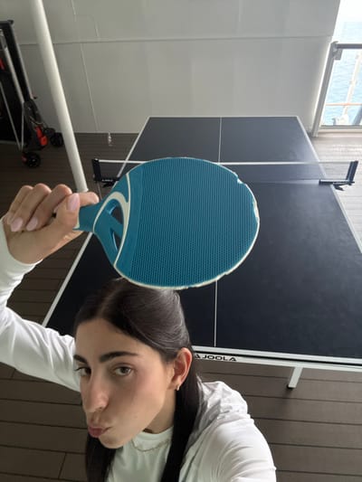 The image displays a close-up, high-angle view of an individual holding a blue and white table tennis paddle. The individual, who has dark hair and wears a white long-sleeved top, looks towards the camera with pursed lips. Their right hand, adorned with light pink nail polish, grasps the paddle handle. The background features a black Joola brand table tennis table with a white net. To the left, a black and orange wheeled device, possibly a scooter, is partially visible next to a white structural pole. The floor is composed of brown planks. Through an opening on the right, a body of water, indicative of the ocean, is visible. The scene is located in the city of Berry Islands, Bahamas.