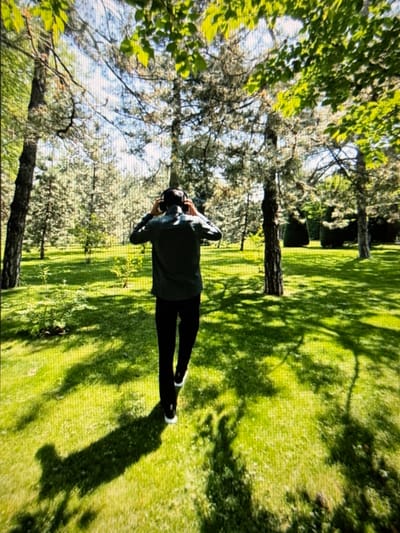 A person is lying on a grassy field under the shade of trees, looking upwards. The individual wears black leggings, white shoes, and a dark-colored top, with their arms crossed behind their head. Sunlight filters through the leaves, creating dappled shadows on the green grass. Trees surround the person, framing the scene, with a clear sky visible through the branches. The composition emphasizes the contrast between light and shadow and the person's relaxed pose.