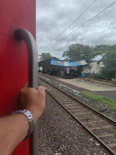 The image shows a view from a moving train at the Dhanora railway station in India. A person's hand, wearing a silver watch, is holding onto a red train car's railing. In the background, the train station platform is visible with a blue and white building labeled "DAGARKHIRI" and also some text in Marathi. There are railway tracks in the foreground and middle ground, with a grassy area and trees to the right. The sky is overcast.