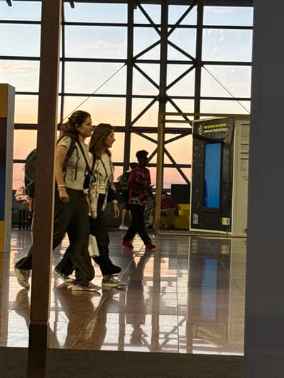The image shows an indoor scene with large windows revealing a sunset sky in Zaventem, Belgium. Two women are walking side-by-side in the foreground, with a person in a red and black patterned shirt walking away from the camera in the middle ground. A man is seated on a yellow structure in the background, and another person is partially visible in the far background. The floor is highly reflective, showing the reflections of people and the window structure. An exhibit with a blue vertical panel is visible to the right of the two women. The overall lighting suggests late afternoon or early evening.