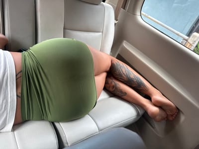 An interior view of a vehicle shows a woman lying across the light grey seats. She wears an olive green skirt and a white top. Tattoos adorn her legs; one features feathers and the other shows a woman's face. Her bare feet rest against the vehicle's side panel near the window. The image is taken from a somewhat elevated angle, focusing on her lower body.