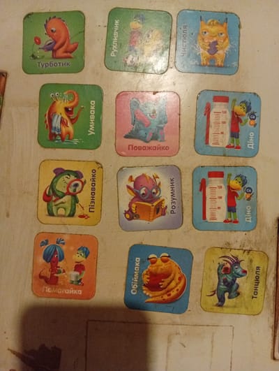 The image displays a collection of small, square, illustrated cards, each featuring a cartoonish monster character. These cards are arranged in a grid-like pattern on a light-colored, textured surface, possibly a table or wall. The cards themselves have rounded corners.
The card in the top left shows a red dinosaur-like creature watering a red flower, with the text "Турботик" below it. To its right, a duck-like creature with yellow and purple plumage is depicted, with the text "Рухливчик" vertically to its left. The next card to the right features a yellow, furry monster with large eyes and small horns, holding a purple object, and the text "Чистюля" vertically to its left.
The second row begins with a green, alien-like creature with large eyes and a blue scarf, and the text "Умивака" vertically to its right. Next to it, a blue, fuzzy monster with large eyes is reading a book, with the text "Поважайко" below it. The third card in this row shows a green dinosaur character standing next to a height chart with markings at 80, 100, and 120, and the text "Дино" vertically to its right. The card also features "Ca" and "D3" icons.
The third row starts with a green creature holding a magnifying glass over a red stain on the ground, with the text "Пізнавайко" vertically to its right. The middle card of this row shows a purple, round monster with glasses reading a book, with the text "Розумник" vertically to its right. The card to its right is similar to the "Дино" card in the row above, featuring a green dinosaur character and a height chart, with the text "Дино" vertically to its right and the "Ca" and "D3" icons.
The bottom row contains three cards. The leftmost card depicts two worm-like creatures, one red with blue pigtails and the other green with a blue tuft of hair, holding plates, with the text "Помагайка" below them. The middle card shows a round, yellow-orange creature with red spots hugging a red object, with the text "Обіймака" vertically to its left. The final card shows a blue-green creature with horns and a red earbud in its ear, dancing, with the text "Танцюля" vertically to its right.
The image does not contain any information about the location in the city of Berezivka, Ukraine. The scene is a close-up of these cards on a surface.