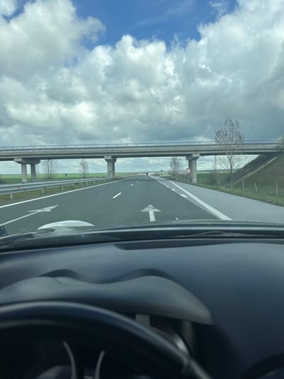 The image shows a view from inside a car on a highway in what appears to be a rural area. To the left of the highway is an elevated railway bridge. The sky is partly cloudy with blue patches. The highway has lane markings and directional arrows. A speed limit sign, presumably "50", is visible. The interior of the car, including the steering wheel, is visible on the right. There are no people visible.