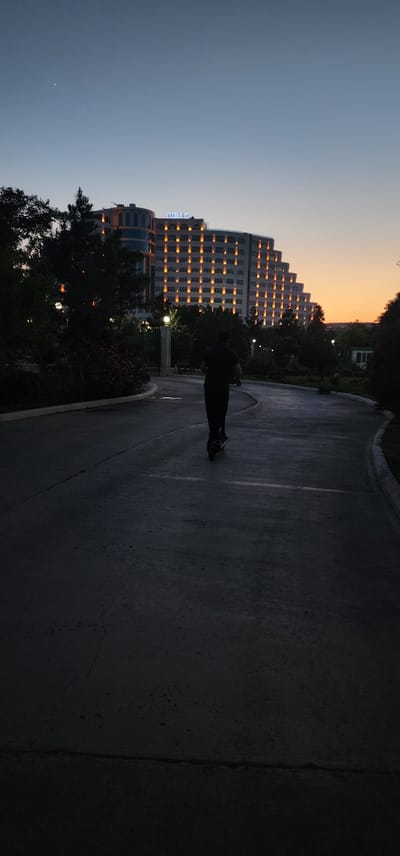 The image shows a silhouette of a person on a scooter against a backdrop of a hotel at dusk. The hotel, labeled "DENIZ," is lit up with warm lights, creating a striking contrast with the darkening sky. The setting appears to be an outdoor park or walkway with a paved path. Trees and some foliage are visible, adding to the natural environment surrounding the urban structure. The time of day is evident from the twilight sky, with hues of orange and blue indicating the transition from day to night. The overall mood is serene and peaceful, with the solitary figure on the scooter adding a dynamic element to the scene. The location is identified as Türkmenbaşy, Turkmenistan.