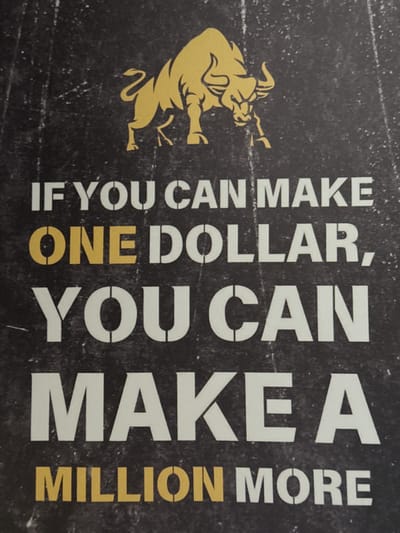 The image displays a motivational quote overlaid on a textured, dark background. The quote, "IF YOU CAN MAKE ONE DOLLAR, YOU CAN MAKE A MILLION MORE," is presented in bold, stenciled letters, with "ONE DOLLAR" highlighted in yellow. Above the text, a stylized golden bull is depicted in a charging stance. The background appears to be a fabric or a wall with a distressed, gritty texture. The image is a still photograph and does not contain any information about a specific location in Oldham, United Kingdom.