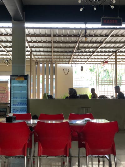 The image depicts the interior of a brightly lit establishment, possibly a cafe or restaurant, with a view towards an outdoor area. In the foreground, four vibrant red plastic chairs with metallic legs surround a glass-topped table, some of which are partially obscured by the chairs. To the left, a glass-door refrigerator is filled with bottled water. In the mid-ground, several individuals are seated behind a low counter. One person, wearing a headscarf, is facing left. Another person, a male in what appears to be a brown uniform, is looking to the left. Further to the right, another man is seated, facing towards the right. Behind them, a lattice-like structure with vertical bars allows light and greenery to filter in from the outside. A sign with red lettering on a black background that reads "KASIR" (Indonesian for Cashier) is visible hanging from the ceiling in the upper right portion of the image. The ceiling is constructed of corrugated metal panels supported by wooden beams. The overall impression is one of a casual, possibly local eatery with a service counter and seating area.