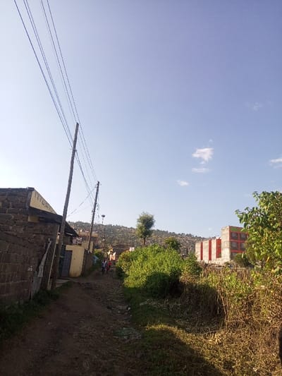 The scene depicts an outdoor environment in Kiamaina ward, Kenya, under a clear blue sky with minimal white cloud cover. An unpaved, uneven dirt path dominates the foreground and extends into the midground. To the immediate left, a stone wall structure is present, behind which parts of a building with a light-colored facade are visible. Two wooden utility poles, positioned diagonally, rise from the left side, supporting multiple overhead power lines that traverse the upper left quadrant of the image. Further along the dirt path in the midground, approximately three to four individuals are observed walking away from the viewer. Dense green vegetation, consisting of bushes and smaller trees, borders the path, particularly on the right side. In the midground, a solitary slender tree stands distinctively. The background features a hillside densely populated with numerous structures, indicative of a residential area. To the far right in the background, a multi-story building under construction or renovation is visible, characterized by prominent red-orange sections.