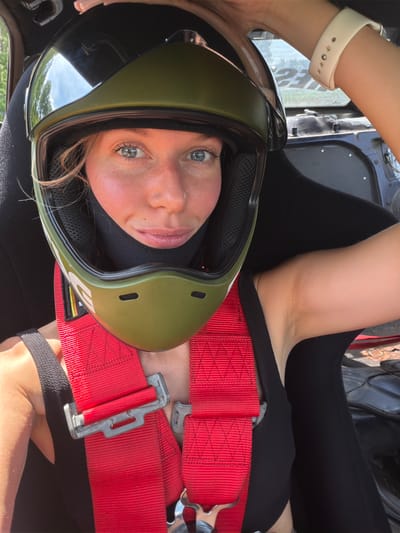 A young woman with blonde hair and blue eyes is sitting in the driver's seat of a race car, wearing a green and black racing helmet and a red racing harness. Her face is partially obscured by the helmet. She is looking directly at the camera with a slight smile. The interior of the car is visible, with black seats and racing equipment. The background appears to be outdoors, with trees and a blurred sign in the distance. The scene is likely set in or around a race track or a location associated with motorsports within the Навадворскі сельскі Савет, Belarus.