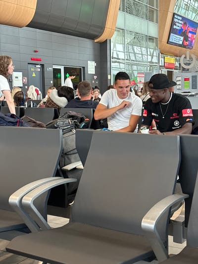 The image depicts a modern airport waiting area. Two young men are seated next to each other in the foreground, facing the viewer's right. The man on the left is wearing a white t-shirt and looking at the man on his right with an open-mouthed expression, as if speaking. The man on the right is wearing a black t-shirt with logos and a black baseball cap, looking down and to his left while holding a can and a white wrapper. They appear to be engaged in a conversation.
Behind them, other passengers are visible, some seated and others standing. Luggage is placed around the seating area. In the background, there are signs and architectural features typical of an airport, including a large digital screen displaying images and text in Russian. The overall environment suggests a public transportation hub, likely an airport. The location is specified as Bolshiye Kabany, Russia.