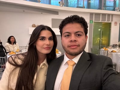 The image captures a selfie-style shot of two individuals in what appears to be a formal indoor event setting, likely a restaurant or banquet hall.
In the foreground, a woman is positioned on the left and a man on the right. The woman has long, dark brown hair, parted down the middle, and is wearing a light-colored (off-white or cream) collared jacket or coat. She has a subtle smile and is looking towards the viewer. The man on the right has dark, curly hair and a short beard or stubble. He is wearing a dark suit jacket, a white collared shirt, and a gold or yellow striped tie. He has a neutral expression and is looking directly at the camera.
In the background, another woman with dark hair is partially visible on the far left, seated at a table and wearing a fluffy white or cream jacket. The room features multiple tables draped with white tablecloths, set with glasses, plates, and yellow napkins. The chairs are silver. The walls are light-colored, and there are some decorative, spherical objects on a shelf in the mid-ground.
The setting includes a large glass partition or window in the upper background, reflecting parts of the interior. Text is visible on this glass, appearing to be "Altea" and "za", possibly part of a word like "Belleza" or "Naturaleza". To the right, there's a green exit sign with an arrow pointing left. Further right, a "No Smoking" sign with a red circle and crossed-out cigarette is visible. The lighting is artificial and bright, suggesting an indoor event that could be taking place during the evening or night. The overall atmosphere appears to be a formal social gathering or celebration.