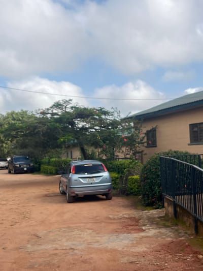 A light blue SUV and a black SUV are parked on a dirt road next to a building with a beige facade. The building has several windows and a dark green roof. A dark metal railing runs along a balcony on the second floor, with green bushes and pink flowers growing beneath it. A large tree with green leaves and some orange flowers stands between the building and the road. The sky above is partly cloudy, with white clouds against a blue background. The scene appears to be outdoors in a rural or semi-rural setting, possibly in Gembu, Nigeria. There is no visible activity or event taking place, and the overall impression is one of a still, daytime scene. The license plate on the light blue SUV is partially visible and appears to be in a Nigerian format.