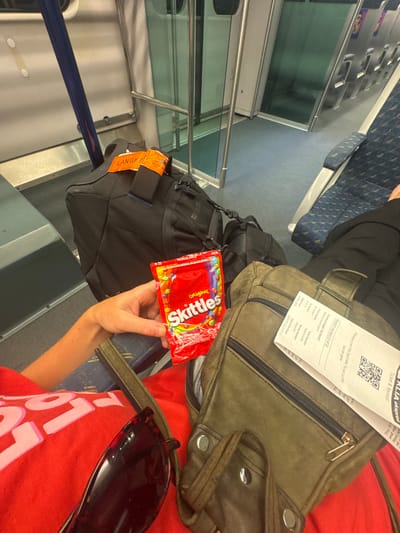 The image displays an interior view aboard a KLIA Ekspres train in Malaysia. A person, seated, has their right arm and upper leg visible, wearing a red shirt with partial white lettering. Their hand holds a red, sealed packet of "Skittles Original" candy. Dark sunglasses with a tortoiseshell frame rest on their lap. Adjacent to the sunglasses, an olive-green leather-like utility bag is positioned, with a white "KLIA ekspres" ticket, dated 2023-01-20 and indicating travel from KL Sentral, protruding from a zipper compartment. On the floor in front of the seated individual, a dark grey or black backpack with an orange luggage tag, partially obscured but showing "LANGKA", is visible. The train's interior features blue patterned fabric seats, grey-blue carpeted flooring, white wall panels, silver vertical handrails, and glass partitions with silver framing. To the far right, a portion of another passenger's leg, clad in dark trousers, is visible in an adjacent seat. The scene is within the transportation network of Malaysia, in the broader region that includes the Kajang Municipal Council.