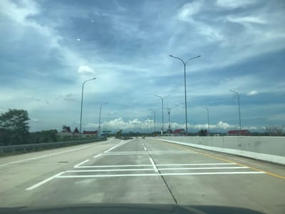 The image shows a view from inside a vehicle driving on a highway in Gohor Lama, Indonesia. The road is gray concrete with white lane markings and a yellow shoulder line. There are several tall streetlights lining both sides of the highway. Buildings with red roofs are visible in the distance. The sky is blue with scattered white clouds.