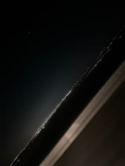 The image displays a nocturnal, distant landscape view of Bogyovtsi, Bulgaria, captured from an elevated perspective. The upper-left quadrant features a dark night sky containing a singular prominent point of light and several smaller, scattered points. A continuous, diagonal line of numerous small, bright artificial lights, consistent with a distant populated area or illuminated roadway, extends across the mid-ground from the lower-left to the upper-right. Below this band of lights, a faint, diffuse glow, indicative of light pollution, is discernible. The foreground, occupying the right side and bottom-right of the frame, consists of a broad, dark, angled structural element, possibly a building roofline or architectural beam, which exhibits a subtle, elongated highlight or reflection along its upper edge. No individuals, direct actions, or interactions are visible.