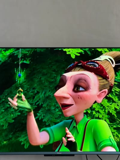 This is a close-up, vertical, animated media file depicting a character from a children's story. The character has pale skin, blonde hair pulled back in a ponytail, and wears red goggles with blue lenses over its eyes. The character is dressed in a vibrant green outfit with red trim. Its face is pale pink, with large, expressive eyes and a small mouth. The character is holding a glowing blue orb with green tendrils extending from it, which appears to be some kind of magical artifact or creature. The character's hand, adorned with red nail polish, is reaching out towards the orb. The background consists of lush green foliage, suggesting an outdoor, natural setting, possibly a forest or a garden. The lighting appears bright, indicating daytime. The overall mood of the image is one of wonder and perhaps a touch of mischief, given the character's animated expression and the glowing object. The word "Haier" is visible on the right side of the screen, indicating the brand of the display device. There are no discernible cues related to Krasnyi Krym, Russia, in the image itself.