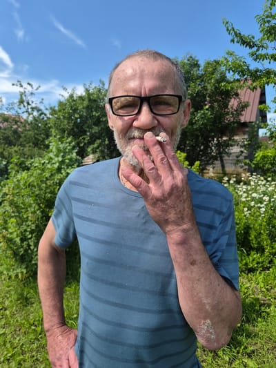 This media file is a portrait of a man in an outdoor setting. He is seen from the chest up, and the image is oriented vertically, with the top of his head to the left. The man has a beard, is wearing glasses with black frames, and is holding a lit cigarette between his fingers, bringing it to his mouth. He is wearing a blue t-shirt with darker blue horizontal stripes.

The background consists of lush green foliage, suggesting a garden or park. There are trees with green leaves and some white flowers. A wooden structure, possibly a building or fence, is visible in the background, partially obscured by the vegetation. The sky above is a clear blue with a few wispy white clouds. The lighting indicates it is daytime, likely a sunny day.

The man's facial expression is neutral, and his focus appears to be on the cigarette. There is no visible text in the image. The overall scene evokes a sense of a relaxed, casual moment outdoors. The location context provided is Novosisoevka, Russia.