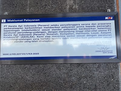 A framed sign is mounted outdoors, likely on a wall or fence. The sign has a blue header with white text that reads "i Maklumat Pelayanan" on the left and the red and white "KAI" logo on the right. Below the header, white text on a light blue background details the commitment of PT Kereta Api Indonesia (Persero) to providing excellent service according to regulations and principles. The bottom of the sign features a registration number "MAK.U/KB.207/VII/1/KA-2023" on the left and a signature, name "DIDIEK HARTANTYO", and NIPP number on the right, dated "Bandung, 27 Juli 2023". The sign is framed in black and mounted against a textured, dark grey surface that appears to be stone or gravel. The location is identified as Desa Tambakreja, Indonesia.