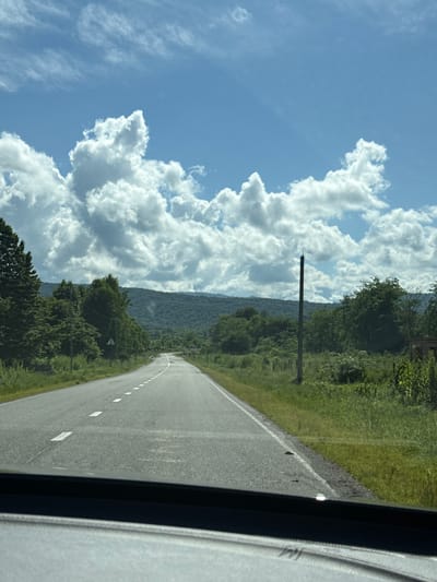 The image depicts a view from inside a vehicle traveling on a paved road in Alagir, Russia. The road is marked with white dashed lines and stretches into the distance towards a tree-covered mountain range. The sky is blue with large, fluffy white clouds. Dense green trees line both sides of the road. On the right side of the road, a tall, dark, slender pole stands prominently in a grassy field, and a section of a brick structure is visible further to the right. The foreground shows the dashboard of the vehicle, partially obscuring the view. The scene suggests a rural or semi-rural setting, possibly a highway or main road leading out of or into Alagir.