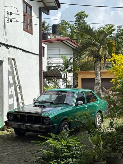 In Kodagoda, Sri Lanka, a vintage, emerald green sedan with a black hood sits parked on a grey paved driveway. The car, bearing the license plate "1255094," is the central focus. To the left, a white building with a brown-trimmed window stands tall, with a silver ladder leaning against its textured wall. Overhead, electrical wires crisscross against a backdrop of a blue sky dotted with white clouds. Behind the car, a tall palm tree dominates the scene, its fronds reaching towards the sky. Further back, a yellow building with a double garage door is visible, partially obscured by lush green foliage and a bright yellow flowering bush. The environment is verdant and tropical, typical of the Sri Lankan landscape.
