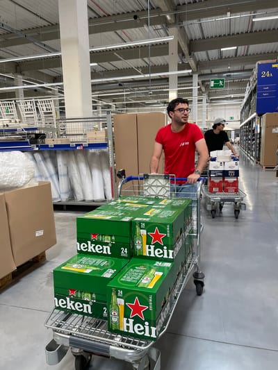 Two men are shopping in a large, well-lit warehouse-style store, identified by a prominent "METRO" sign on a blue banner hanging above an aisle. The store features high ceilings with exposed metal structures, fluorescent lighting, and extensive shelving stocked with various products.
In the foreground, a man wearing a red t-shirt with "IED" printed on it and glasses is pushing a large metal shopping cart. His cart is filled with several green cases of Heineken beer, each prominently labeled "Heineken ORIGINAL" and indicating "24 33cl BOTTLES". He looks forward with a calm expression.
Further down the aisle, another man, dressed in a black t-shirt and a white baseball cap, is pushing a second shopping cart. His cart contains multiple cases of Corona Extra beer, marked "24", and boxes of Campari, with "CAMPARI" and the number "426840" visible.
The shelves along the aisle hold numerous cardboard boxes and white-wrapped items. A green emergency exit sign with a running person symbol is visible on a pillar. The floor is a light gray, industrial finish. The scene depicts a bulk shopping trip in a cash-and-carry environment, characteristic of a Metro store. The specified location is Su Masu/Elmas, Italy.