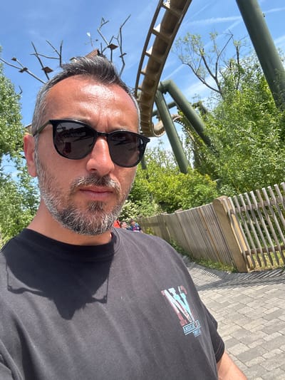 A selfie of a middle-aged man wearing sunglasses and a black t-shirt. He is outdoors with trees and a light-colored rollercoaster track visible in the background. There is a wooden fence and stone pavers near him. Based on the rollercoaster track and general environment, the photo appears to have been taken in Erlebnispark Tripsdrill, located in Cleebronn, Germany.