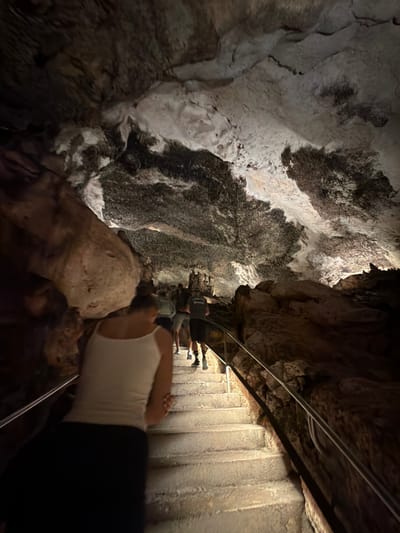The image shows the inside of the Caves of Drach in Manacor, Spain. The view is from a low angle looking upwards towards a rock cave wall on the left and a stairway and cave ceiling on the right. A person is in the foreground, partially visible, wearing a white tank top and black leggings. Several other people are walking down the stairway in the background. The environment is dark and illuminated by artificial lights.