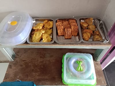 Three metal trays of fried food rest on a shelf. One tray holds battered and fried vegetables, the middle tray contains fried tempeh, and the last tray holds fried tofu. Tongs sit alongside the tofu. A plastic food cover with a yellow handle sits to the left of the trays. Below, on a lower shelf, sits a green and transparent plastic container, also with a lid.