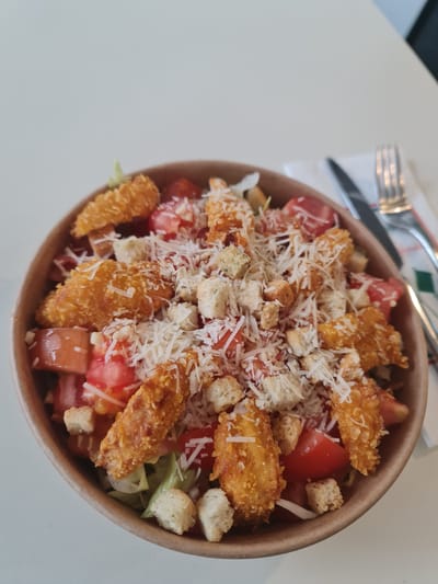 This close-up image presents a vibrant salad served in a disposable, brown paper or cardboard bowl, positioned on a light-colored, possibly white, table or countertop. To the left of the bowl, a silver fork and knife rest neatly on a white napkin adorned with a subtle green and red patterned border.
The salad itself is generously portioned and layered with various ingredients. Several pieces of golden-brown, breaded fried chicken are prominently featured across the top. Mixed with the chicken are numerous bright red, diced tomatoes and light brown croutons, adding a crunchy texture. A copious amount of white, grated cheese is sprinkled over the entire salad, creating a visually appealing topping. Beneath these components, green leafy lettuce forms the base of the salad.
The scene depicts a meal prepared for consumption, likely in a casual dining environment or as a takeout order. The lighting is bright and even, suggesting an indoor setting during the daytime or in a well-lit interior. No visible text or branding is present in the image.