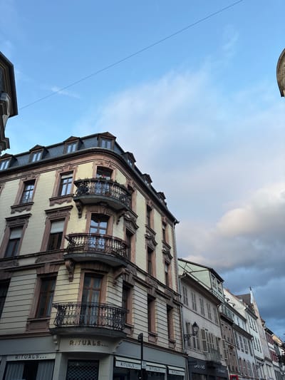 This is a low-angle, eye-level shot of buildings in Colmar, France, with a focus on a corner building. The sky is partly cloudy with a mix of blue sky and dark, dramatic clouds. A thin, dark wire is visible stretching across the sky.

The main building is a multi-story structure with a light yellow facade and ornate brown trim around the windows and along the roofline. It features several windows with brown wooden frames, some with closed white shutters. Two decorative balconies with black wrought-iron railings protrude from the corner. The balconies have planters with flowers. The uppermost part of the building has a dark gray mansard roof with dormer windows.

Below the balconies, the corner building has a storefront with "RITUALS..." signage in gold letters. The shop appears to be a retail establishment, with displays visible through the windows. Other storefronts, including "promod," are visible along the street to the right, with their awnings and signs. The architecture of the adjacent buildings is varied, with shutters and different colored facades, typical of European city streets. The overall impression is of a commercial street in a historic European town.