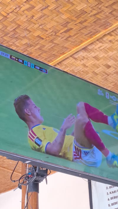 This is a media file showing a close-up, angled view of a television screen displaying a soccer match. The screen is mounted on a wooden beam structure.
The subjects on the screen are two soccer players in uniform. One player, in the foreground, is wearing a yellow jersey with blue trim and red accents on the sleeves. They appear to be looking upwards. Another player, in the background, is wearing a white jersey with red accents and red sleeves. The game is being played on a green field, and the scoreboard indicates "COL 0 - 0 COD" with a time of "37:37".
Visible text on the screen includes "COL", "COD", "37:37", and several lines of text on a white background to the right of the game which appear to be a list, possibly of fouls or player statistics, with entries like "2. BEB...", "3. KARIB...", "4. BEBE...", and "5. BEBE...".
The setting is indoors, suggested by the television screen and the surrounding wooden structure, possibly a covered outdoor area given the natural materials. The overall lighting appears to be natural, possibly daylight. The camera perspective is from below and to the side of the television.