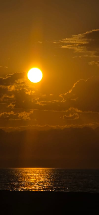 A photograph captures a sunset over a body of water, likely a sea or large lake, with a dark foreground suggesting a beach or land. The sun, a brilliant white orb, is positioned in the upper left quadrant, partially obscured by wispy, golden clouds. Rays of light fan out from the sun, creating a dramatic atmospheric effect. The water reflects the intense golden light of the sunset, creating a shimmering, almost sparkling surface. The horizon line is clearly defined, separating the water from the hazy sky. The overall color palette is dominated by warm oranges and yellows, indicative of a sunset. The image does not contain any people or specific urban landmarks that would identify it as being in Basbeshik, Kyrgyzstan.