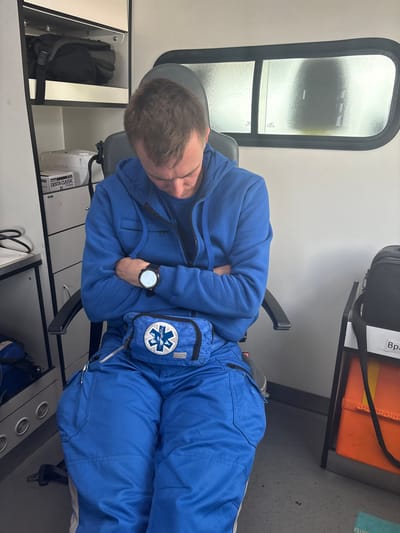 A young man wearing a blue paramedic uniform with a medical symbol patch on his waist bag is seated in a chair inside what appears to be an ambulance. He is asleep, with his head bowed and arms crossed. The background shows medical supplies and equipment, including shelves with bags and boxes, and a window with a frosted appearance. The overall scene suggests a moment of rest during or after a duty shift.