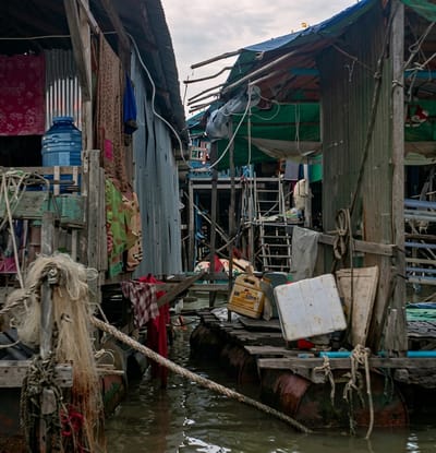 The image depicts a densely packed slum on a body of water, likely a river or lake, in Soutr Nikom, Cambodia. The structures are haphazardly constructed from salvaged materials, including corrugated metal, wood, and tarpaulin, with many appearing to be floating or built on stilts. Laundry hangs from lines strung between buildings. A large blue water jug is visible on a wooden platform. In the foreground, a boat or raft is tied up with thick ropes, and fishing nets are draped over its side. Several barrels are visible beneath a wooden platform, suggesting a floating structure. A yellow plastic container and a white plastic cooler are placed on this platform. The water is murky and appears to be polluted. The overall scene conveys a sense of poverty and makeshift living conditions.