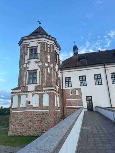 The image shows a close-up view of the Mir Castle Complex, located in the village of Mir, within the Karelichy District of Grodno Region, Belarus. The castle features a prominent octagonal tower constructed of red brick and stone, adorned with decorative white elements. The tower has multiple windows, some with arched tops, and is capped by a tiled roof with a weather vane. Adjacent to the tower is a white building with a dark tiled roof and several dark-framed windows. A paved walkway with white railings leads towards a dark wooden door on the white building. The ground surrounding the castle is covered in green grass. The sky above is blue with scattered white clouds.