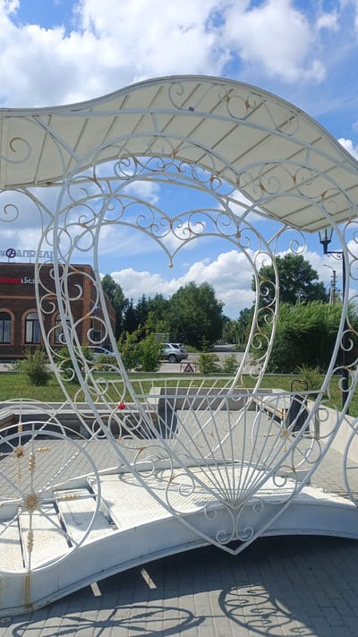 A white metal gazebo, shaped like a heart, is the central subject of this outdoor photograph. The gazebo features intricate scrollwork and a white canopy. Through the heart-shaped opening, a brick building with arched windows and signage is visible. The text on the building reads "АПРЕД" and "КрасноярскБе". Beyond the building, a street is visible with passing cars and lush green trees. The scene is set against a bright blue sky with fluffy white clouds, suggesting a sunny day. The time of day appears to be midday, judging by the shadows cast by the gazebo. The overall impression is a peaceful, possibly recreational setting, with the gazebo serving as a decorative element in a public space.