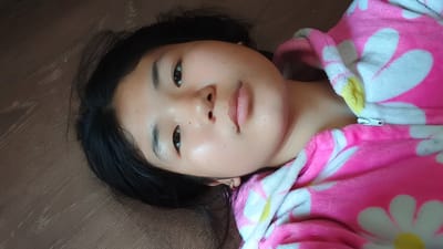 A young Asian girl with black hair lies on her back on a brown, wood-grain textured surface. She is wearing a pink fleece robe with white and yellow daisy patterns. Her head is slightly tilted to her left. The girl's face is illuminated from above, with her eyes open and looking upwards. Her skin appears smooth with subtle blemishes and a dewy sheen. Her lips are closed and slightly parted. A small gold earring is visible in her left ear. The robe is zipped up at the front, with a white zipper pull. The lighting suggests an indoor setting, likely in the city of Uchkun, Kyrgyzstan.