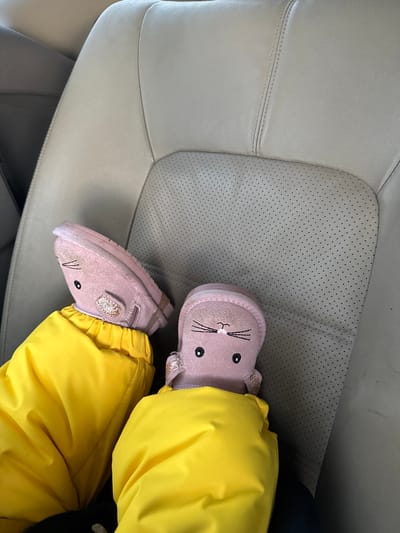 The image displays the lower limbs of an individual, likely a child, situated within a vehicle in Bilimbay, Russia. The individual is wearing bright yellow, possibly padded or waterproof, pants. Their feet are clad in light pink, animal-themed boots, which feature embroidered black eyes, whiskers, and a small pink nose, resembling rabbits or mice. A light-colored, glitter-accented strap is visible on the side of the left boot near the ankle. The feet are oriented upwards, resting against a light-colored, likely beige or light grey, leather car seat. The car seat exhibits perforated sections on its lower central area and distinct stitching along its seams, indicative of an automotive interior.
