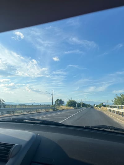 The image captures a view from inside a vehicle, looking out through the windshield at a two-lane asphalt road stretching into the distance under a blue sky with scattered white clouds. The road has white lane markings. On the left side of the road, a metal guardrail runs alongside, with dry vegetation and trees beyond it. Overhead wires for what appears to be an electric train or tram system are visible. On the right side of the road, there is a similar guardrail, a roadside sign with blue and white markings, and more dry vegetation and trees. In the far distance, mountains are visible on the horizon. The foreground shows the dashboard of the vehicle. The scene is consistent with a rural or suburban area, potentially on the outskirts of Graf Ignatievo, Bulgaria, given the presence of infrastructure like overhead wires and guardrails that might be found along a main road. There are no people visible in the image.