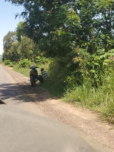 A person wearing a striped shirt and a cap is seated next to a parked motorcycle on the side of a dirt road. They are positioned in the shade of overgrown green vegetation, with trees and foliage visible in the background. The road appears to be a rural or semi-urban area, with a hint of more trees in the distance. The scene suggests a pause or rest during a journey. The location is identified as Mangu, Nigeria.