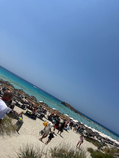 The image depicts a bustling beach scene in Ayia Napa, Cyprus. The sand is light-colored and stretches towards the turquoise waters of the Mediterranean Sea. Numerous beach umbrellas with thatched roofs and white canopies are set up, providing shade for beach chairs and loungers occupied by people. The sea is populated with swimmers, and several watercraft, including what appears to be a jet ski, are visible. In the foreground, people are walking along the sandy shore, some carrying bags and one man is holding a yellow toy. The sky is a clear, vibrant blue, indicating a sunny day. The overall impression is one of a popular tourist destination during peak season.
