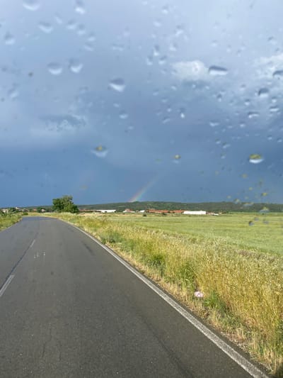 The image shows a view from inside a vehicle, looking out through a rain-streaked windshield. Outside, there is a two-lane asphalt road curving to the right. To the right of the road is a field of tall grass and vegetation. Beyond the field is a sparse settlement. In the background, a segment of a rainbow is visible in the sky. The image was likely taken in Belovitsa, Bulgaria.