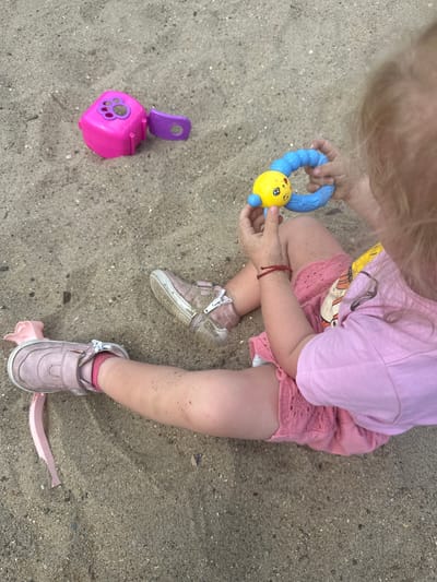 A young child, with blonde hair visible, is seated on a sandy surface, likely a beach or playground. The child is wearing a pink t-shirt and pink lace shorts, and pink shoes. They are holding a blue and yellow toy, a rattle-like object with a face on the yellow part. To the left of the child, there are two pink and purple plastic toys, possibly sand molds or building blocks, lying on the sand. The sandy environment is visible throughout the image, with some small pebbles and debris scattered on the surface. The scene does not contain any information that can determine the location in the city of Donchovci, Bulgaria.