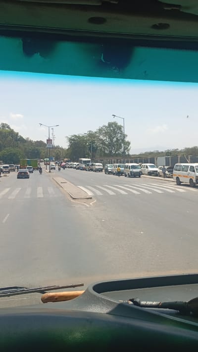 The image captures a forward-facing view from the interior of a vehicle, looking out onto a multi-lane asphalt road under a clear sky in Viwandani ward, Kenya. The vehicle's dark-colored dashboard is prominent in the lower foreground, displaying a light brown, wooden, bat-like object resting horizontally and, to its right, a dark bundled object with a wrapped handle. The windshield's upper portion features a blue-green tint with visible wear and dark spots.
The external scene shows a wide road with a low, narrow concrete median separating traffic lanes. Two distinct pedestrian crosswalks are marked across the visible lanes. On the left side of the road, traffic is present, including a dark sedan, a white van, a green truck, and two motorcycles. On the right side, approximately 15-20 vehicles, predominantly white passenger vans (matatus) and one larger white bus, are parked or stopped in a queue extending into the distance. One individual in a yellow shirt is visible near the parked vehicles on the right. Multiple streetlights with single arms line the road. In the background, dense green foliage, a treeline on a hill to the left, and distant hills are visible on the horizon. A low fence or wall runs parallel to the parked vehicles on the right side of the road.