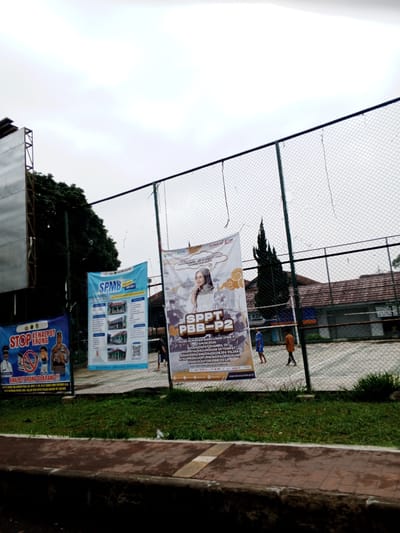 The image captures an outdoor scene, likely near a sports court, indicated by the fencing and a blurred court surface in the background. The sky is overcast and appears grey. In the foreground, there is a grassy area and a textured walkway. Prominently displayed are three large banners attached to the fence.

The leftmost banner, predominantly blue, features text and images related to "STOP KENALPOT BRONG". It depicts a uniformed officer, a child holding what appears to be a toy, and a symbol of a crossed-out exhaust pipe, suggesting a campaign against noisy motorcycle exhausts.

The central banner, a lighter blue and white, displays the acronym "SPMB" and includes images of buildings and text that appears to be informational, possibly related to an academic or administrative process.

The rightmost banner, white with orange accents, features a woman wearing a hijab and text that reads "SPPT PBB-P2". This banner seems to be an announcement or information about property tax.

In the background, beyond the banners and fence, there are buildings with brown roofs and trees. A few people are visible on the sports court, one wearing a blue shirt and another in an orange shirt, seemingly engaged in activities on the court. The overall impression is a public space with educational or civic announcements displayed. The location is not Pelwehera, Sri Lanka.