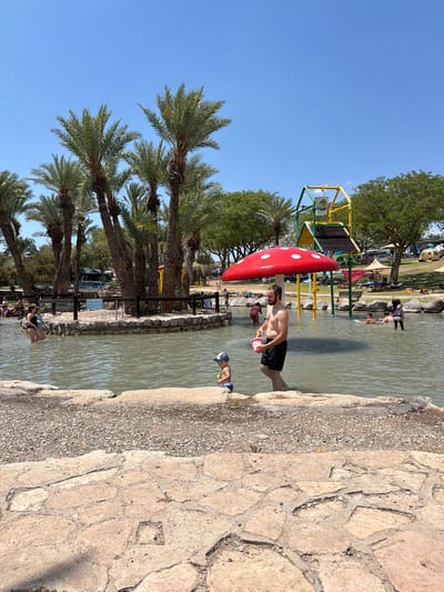 The image depicts a recreational scene in Emek HaMaayanot Regional Council, Israel. A man wearing black shorts stands in shallow water, holding a pink bucket and interacting with a child in a blue and white cap. Adjacent to them is a structure resembling a large red mushroom with white spots. In the background, there is a playground structure with yellow and green elements. Palm trees line the area to the left of the frame, while the edge of the water meets a stone pathway on the right. Other individuals are present in the water and on the grassy area further in the background. The sky is clear and blue.