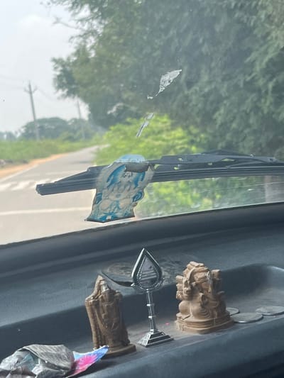 The scene is observed from inside a vehicle located in Srivaikuntam, India, looking forward through the windshield. The foreground displays the vehicle's dark grey dashboard, which holds several items. From left to right, a crumpled wrapper with blue and pink elements rests beside a brown, vertical figurine resembling a robed deity or sage. To its right, a silver-colored, stylized lamp stand features a teardrop-shaped top with dark markings, potentially representing a Shiva Lingam. Further right, a brown figurine of Lord Ganesha, depicted seated with multiple arms, is positioned next to several silver-colored Indian rupee coins. The dashboard surface exhibits a fine layer of dust.

Above the dashboard, the windshield is visible, showing streaks, dirt, and several white, irregular patches, possibly remnants of old adhesive or damage. A single black windshield wiper blade extends across the lower portion of the glass. Partially obscuring the wiper is a torn, blue and white sticker depicting a Hindu deity.

Beyond the windshield, the exterior environment unfolds. A paved road, light grey in color, curves towards the left. Dense, green foliage lines both sides of the road, indicating a vegetated area. Multiple utility poles with associated power lines are visible on the left side of the road, extending into the background. The sky appears overcast or hazy. No individuals or active vehicles are present in the exterior view.