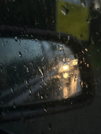 The image is a close-up view through a wet surface, likely a window or windshield, with raindrops distorting the view. In the background, there are two bright, circular light sources that appear blurred and diffused by the water droplets. To the left of the lights, there is a muted, yellow-green rectangular shape, possibly a sign or a colored object viewed through the rain. A dark, curved object, resembling a car's side mirror or a portion of a vehicle's interior, is visible behind the lights and to the right. The overall scene is dark and appears to be set at dusk or night, as indicated by the artificial lights. The presence of raindrops suggests it is raining. The location context provided is Pavlovskii, Russia, but there are no specific visual cues within the image to confirm this location. The mood evoked is somewhat abstract and perhaps melancholic due to the rainy weather and the blurred, obscured view. No text is visible in the image.