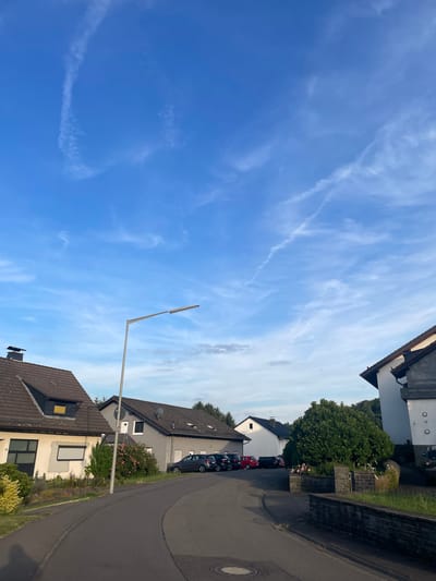 The image depicts a quiet residential street in Wiehl, Germany, under a bright blue sky with wispy white clouds and contrails. A paved road curves through the scene, lined with several houses of varying architectural styles. On the left, a light-colored house with a dark-tiled roof and a dormer window is visible. To its right, a grey house with a satellite dish is situated, with a small rose bush in front. Further down the street, more houses and parked cars, including a dark grey hatchback and a red car, are present. A tall, modern street lamp with a single light fixture stands prominently in the middle ground, casting no discernible shadow. A dense green tree dominates the right side of the frame, with a low brick wall running along the edge of the street in front of it. The overall impression is one of a peaceful suburban environment during daylight hours.