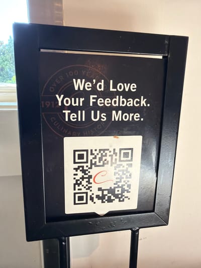 A black framed sign with white text reads "We'd Love Your Feedback. Tell Us More." Below the text is a QR code. The sign is positioned in what appears to be an indoor setting, possibly a restaurant or establishment that values customer feedback. The background suggests a wall and a window with greenery visible outside. The sign itself is mounted on a black metal stand.